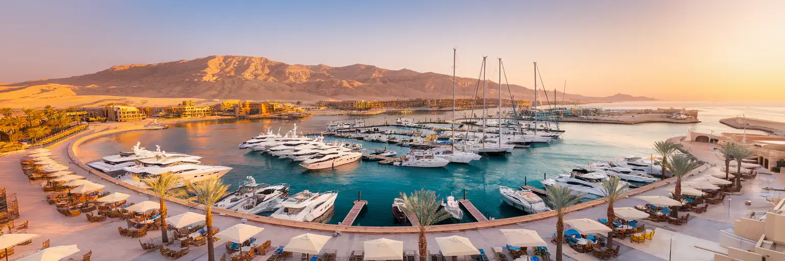Hurghada Marina at golden hour with white yachts moored along the palm-lined waterfront promenade and desert mountains in the background
