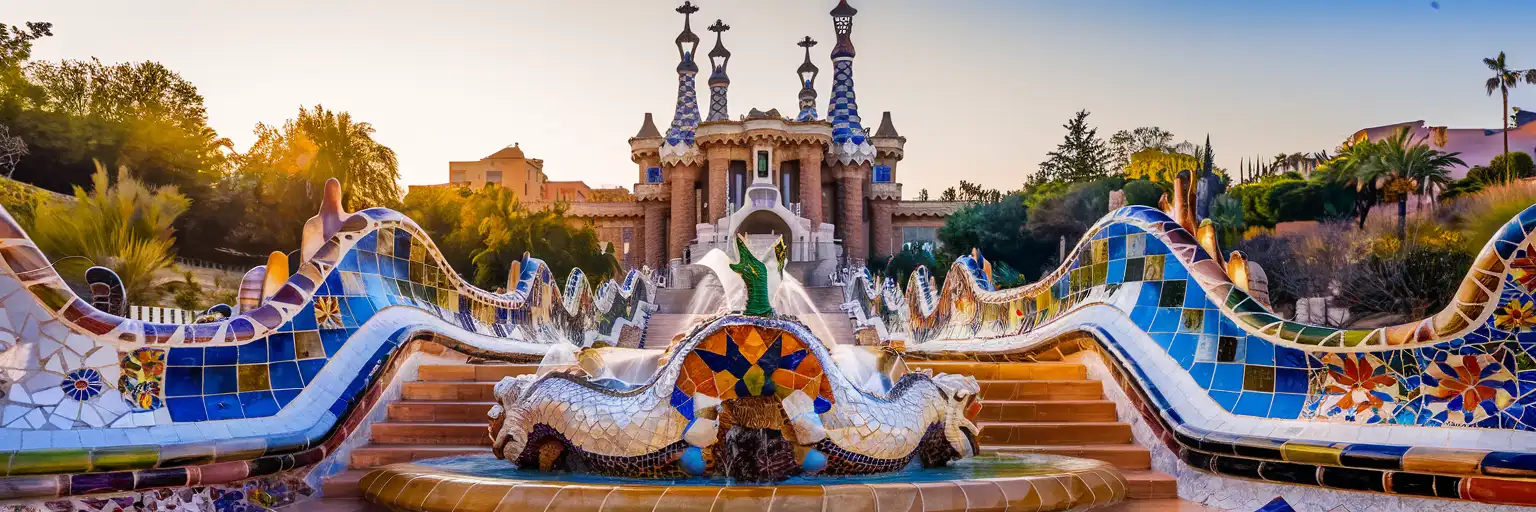 The mosaic dragon fountain at Park Güell's main entrance with twin gatehouses and Mediterranean sky at golden hour