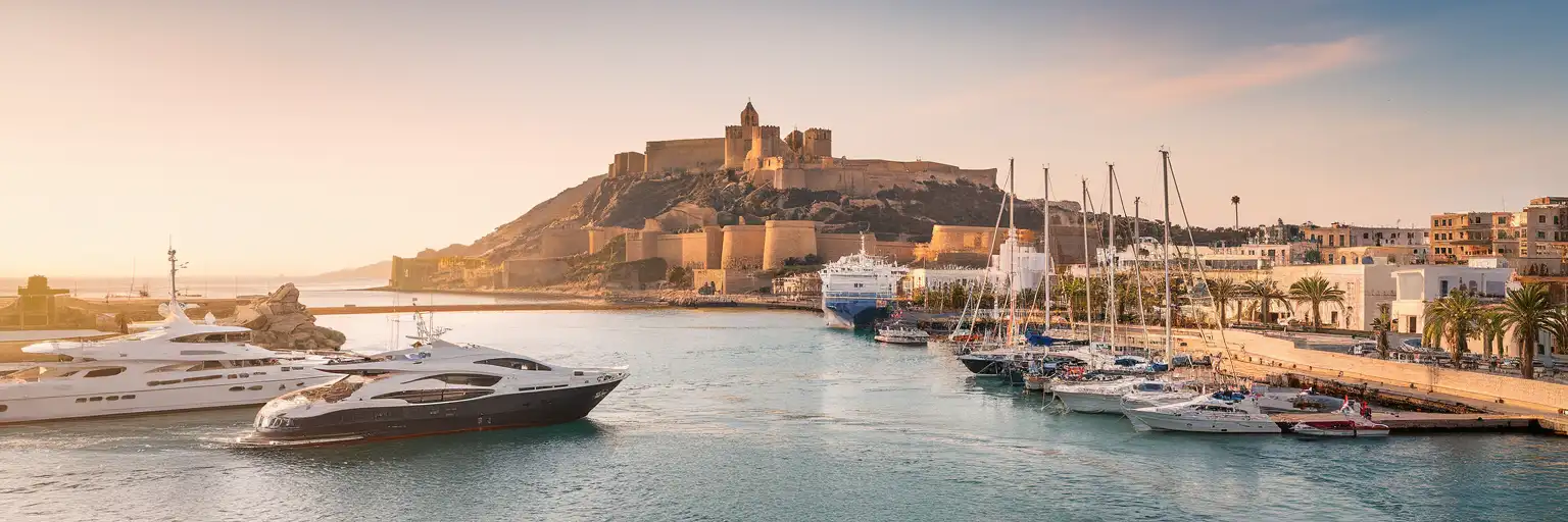 Wide golden-hour exterior view of Port of Ibiza with ferry quays, marina waterfront, and Dalt Vila above the harbor