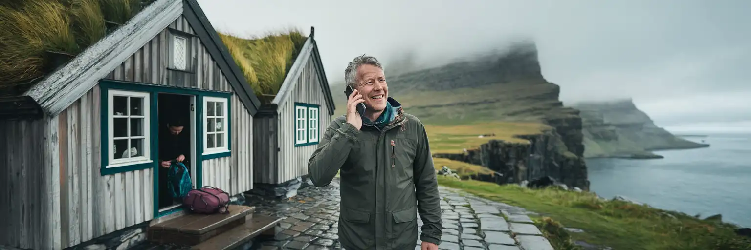 Traveller making a phone call outside a traditional Faroese grass-roofed house with dramatic fjord cliffs in the misty background