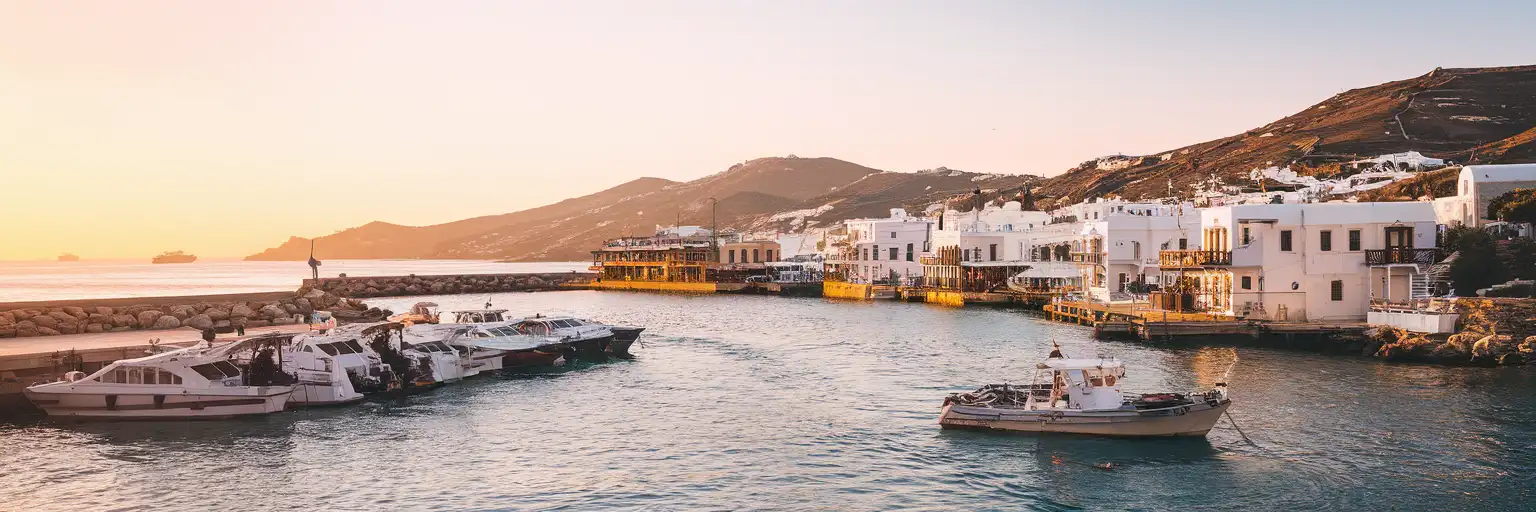 Wide sunset view of the Old Port of Mykonos waterfront with ferries, cruise tenders, and whitewashed harbor buildings