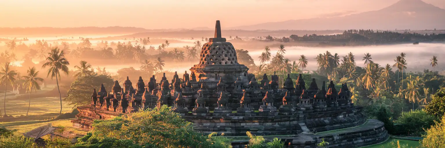 Borobudur temple complex rising from tropical jungle at golden hour with volcanic mountains in the misty distance, Central Java, Indonesia