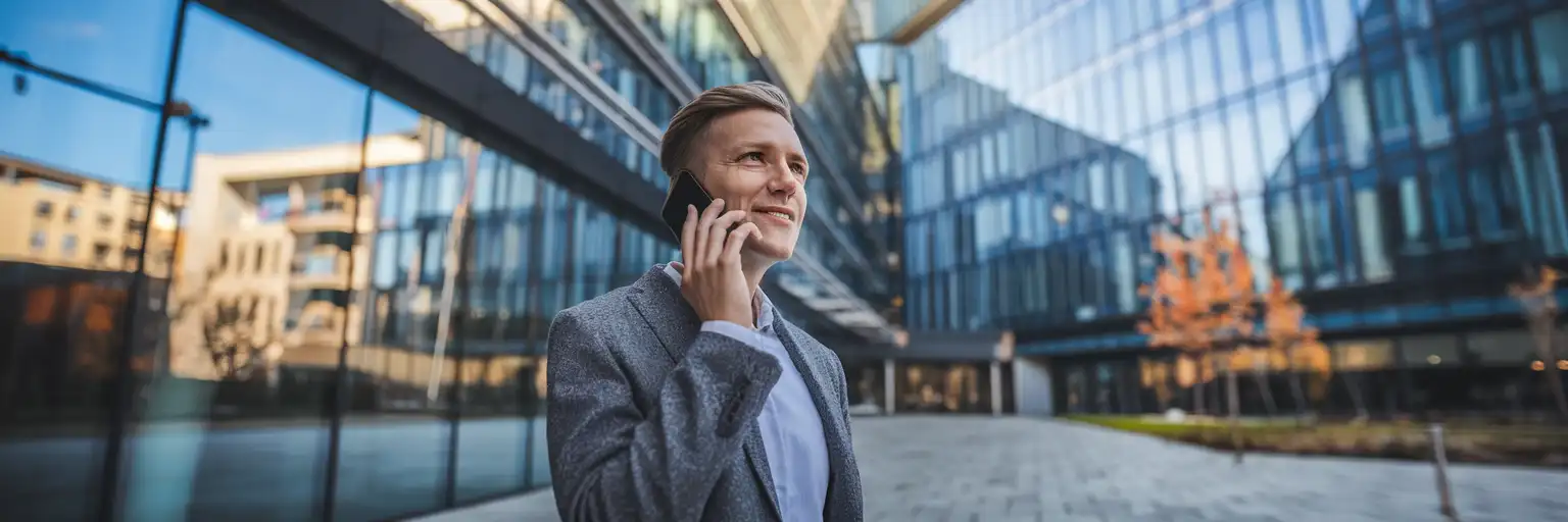 Profesjonalny podróżnik rozmawiający przez telefon przed nowoczesnym budynkiem biznesowym w Luksemburgu podczas jesiennego popołudnia