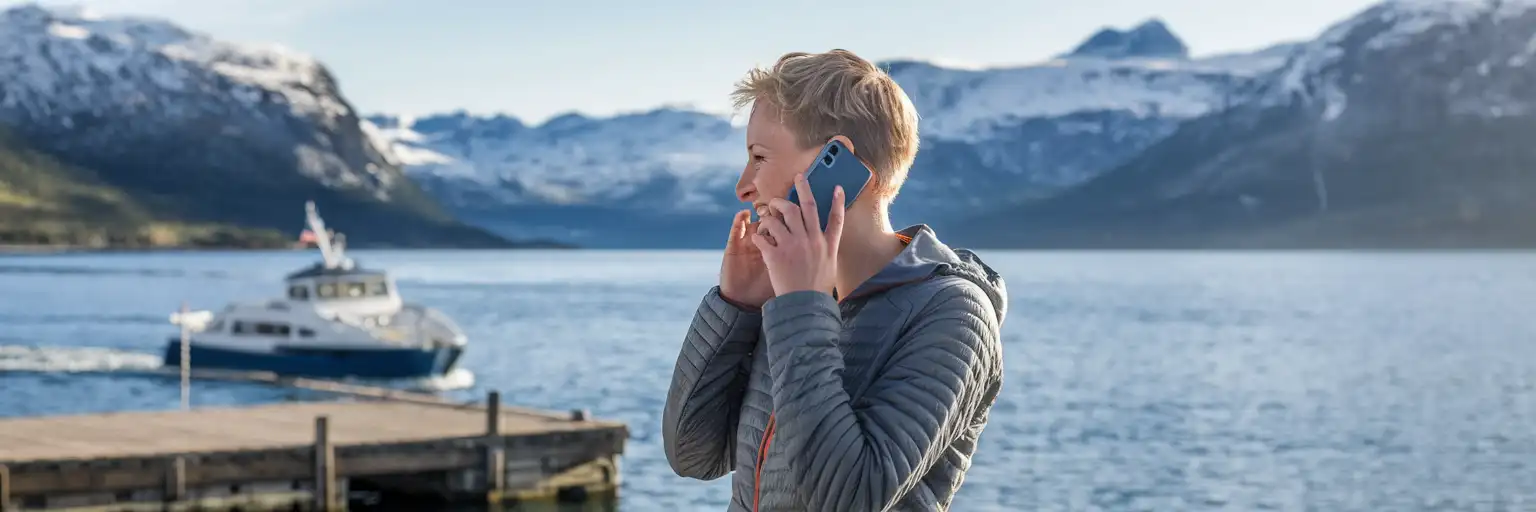 Norveç fiyordunun yanında, ahşap bir iskelede telefonla konuşan bir gezgin, arka planda dağlar