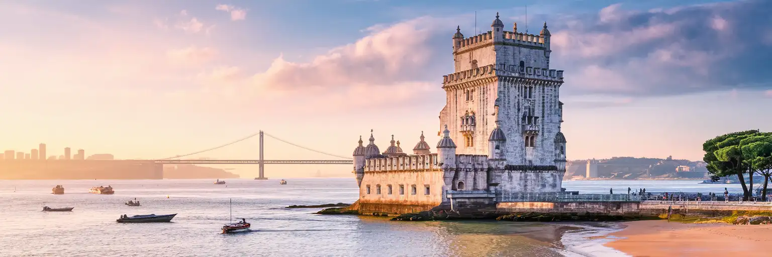 Belém Tower standing in the Tagus River at golden hour with the 25 de Abril Bridge visible in the distance across the Lisbon waterfront
