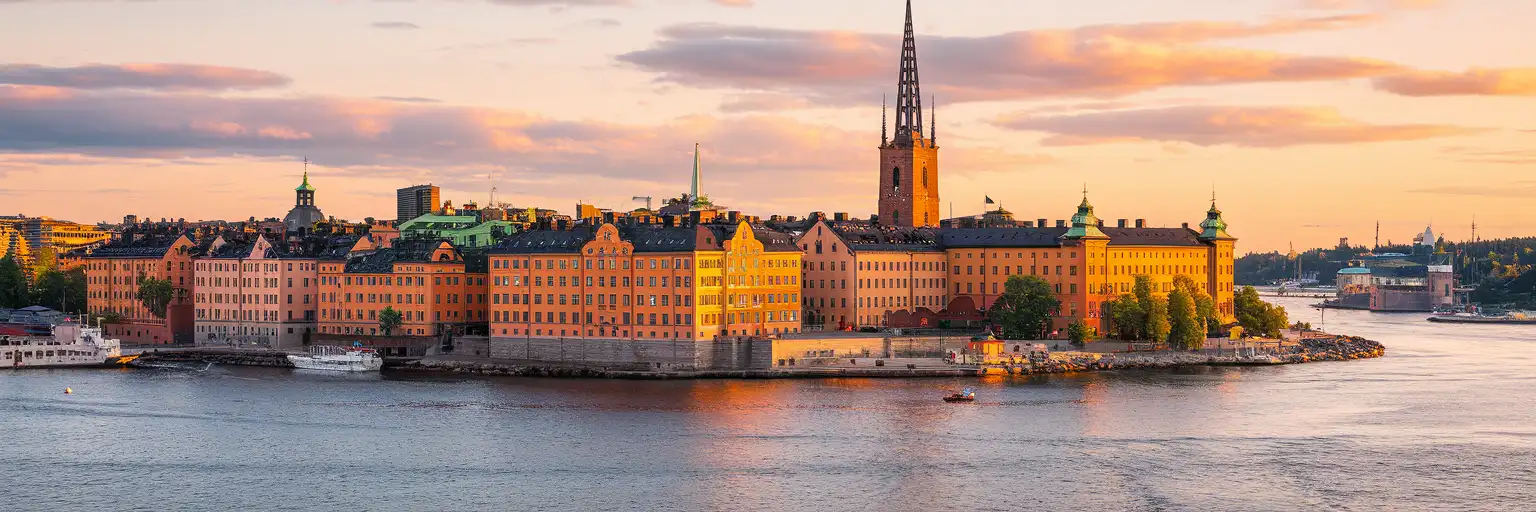 Faleza Gamla Stan din Stockholm cu clădiri medievale portocalii și turla Bisericii Riddarholmen reflectate în Riddarfjärden la ora de aur