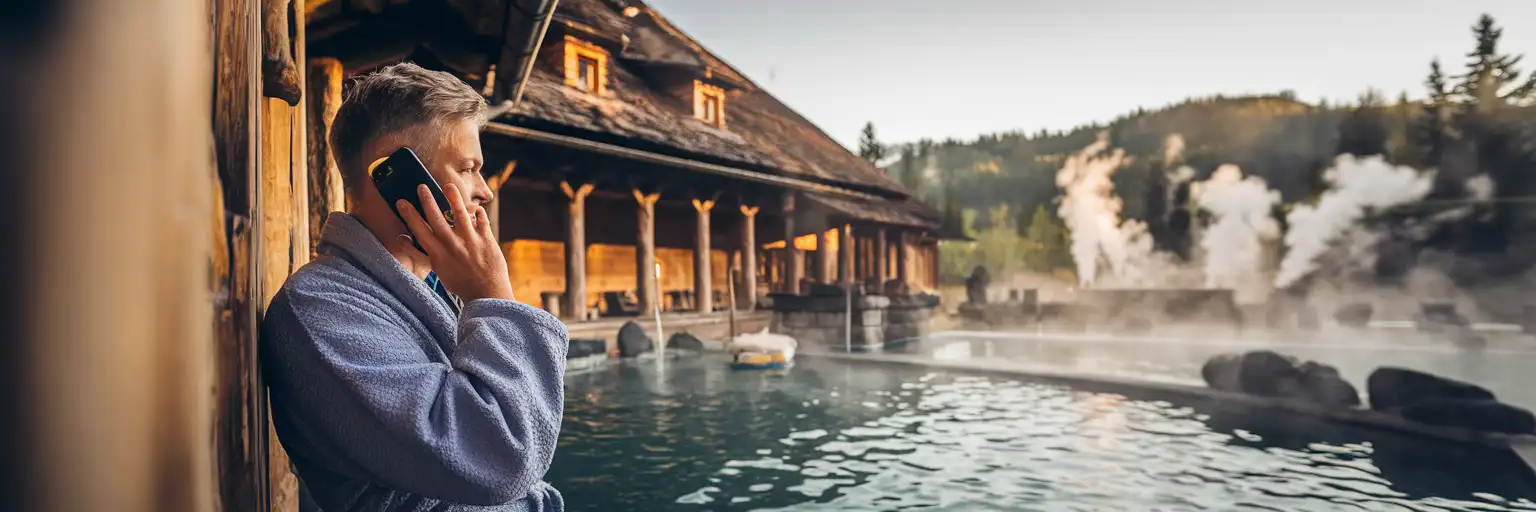 Podróżnik odbierający telefon przed słowackim spa termalnym z parą unoszącą się nad basenami i Karpatami w tle