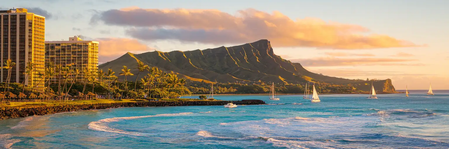 Diamond Head crater rising above Waikiki Beach at golden hour with sailboats dotting the turquoise Pacific waters