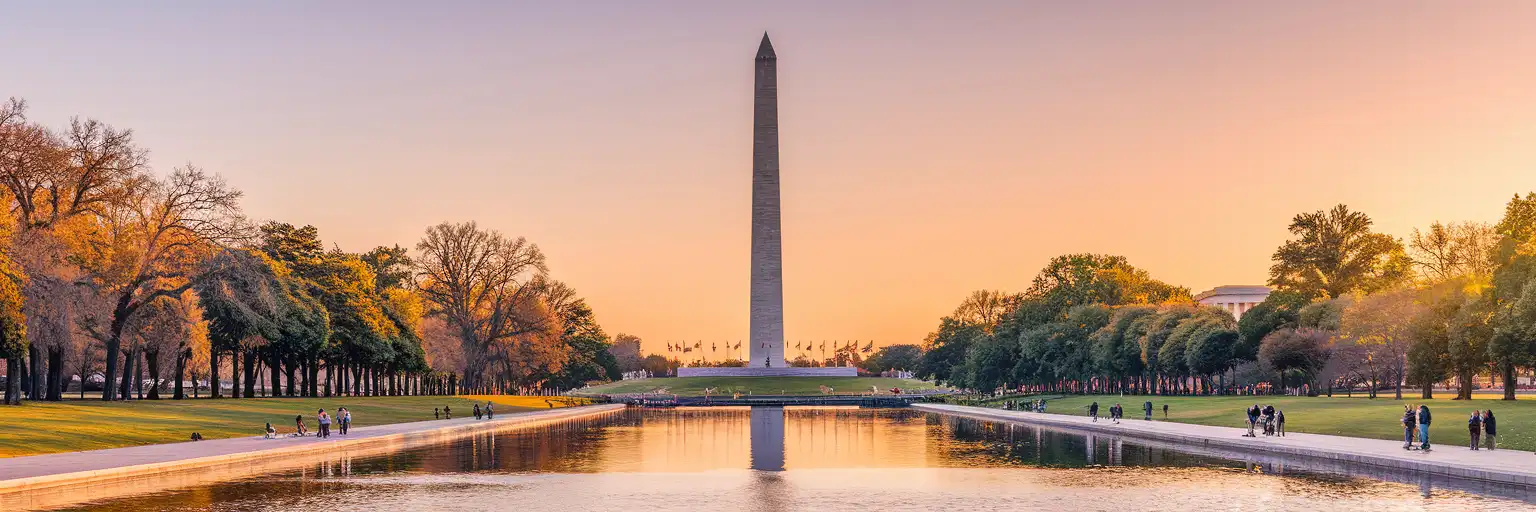 Washington Monument obelisk rising above the National Mall Reflecting Pool at golden hour with American flags and Lincoln Memorial in distance